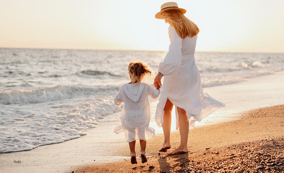 a woman and a young girl wearing white dresses and holding hands on the beach.